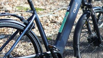 Un vélo électrique noir, avec une marque bien visible et une fourche avant à suspension, est garé sur un chemin de gravier bordé d’arbres.