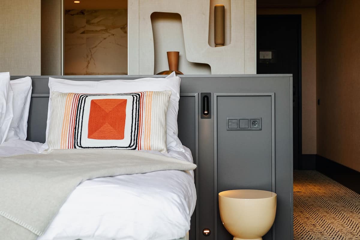 Une chambre d’hôtel avec un lit, une table de chevet, une lampe fixée au mur et une moquette à motifs.