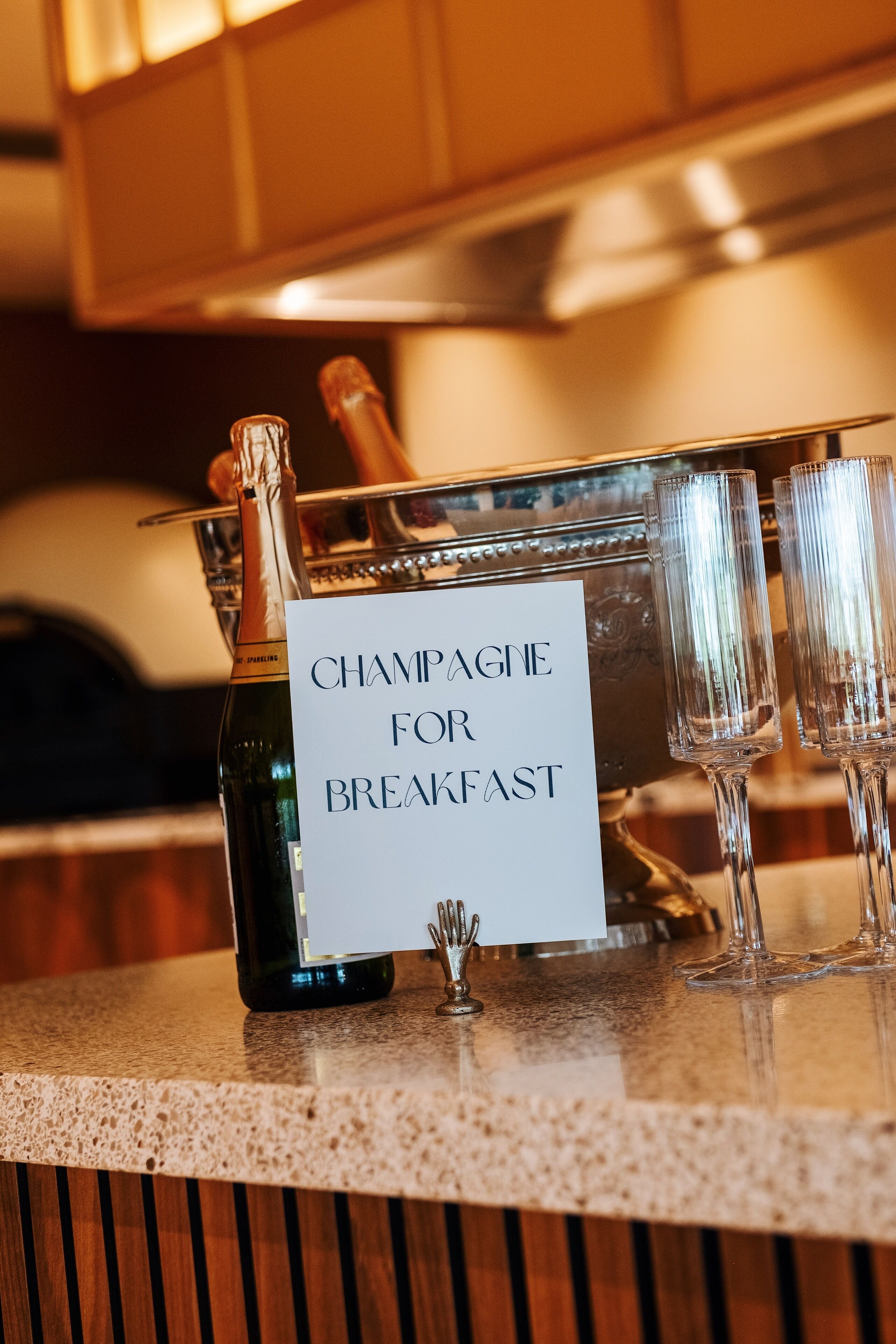 Une bouteille de champagne et deux verres posés sur un comptoir, avec une pancarte indiquant : « Champagne au petit-déjeuner ».