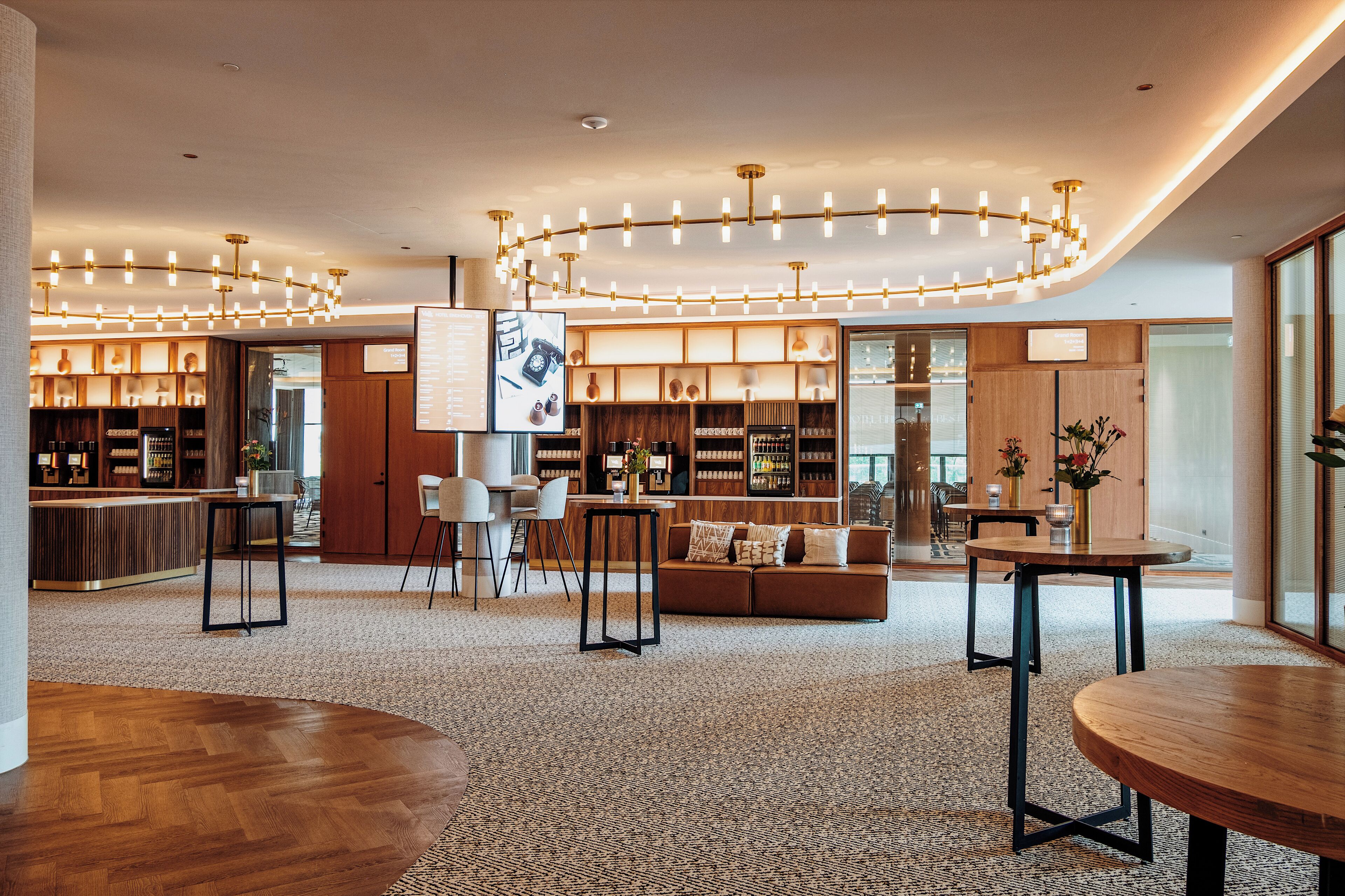 Een moderne hotel lobby met een houten receptiebalie, een zithoek met een bank en tafels, en een bar met flessen op de planken.