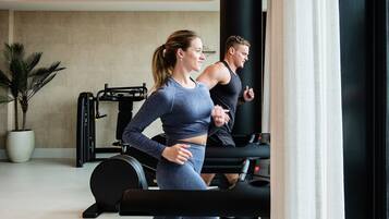 Two people running on treadmills in a gym with exercise equipment and a potted plant in the background.