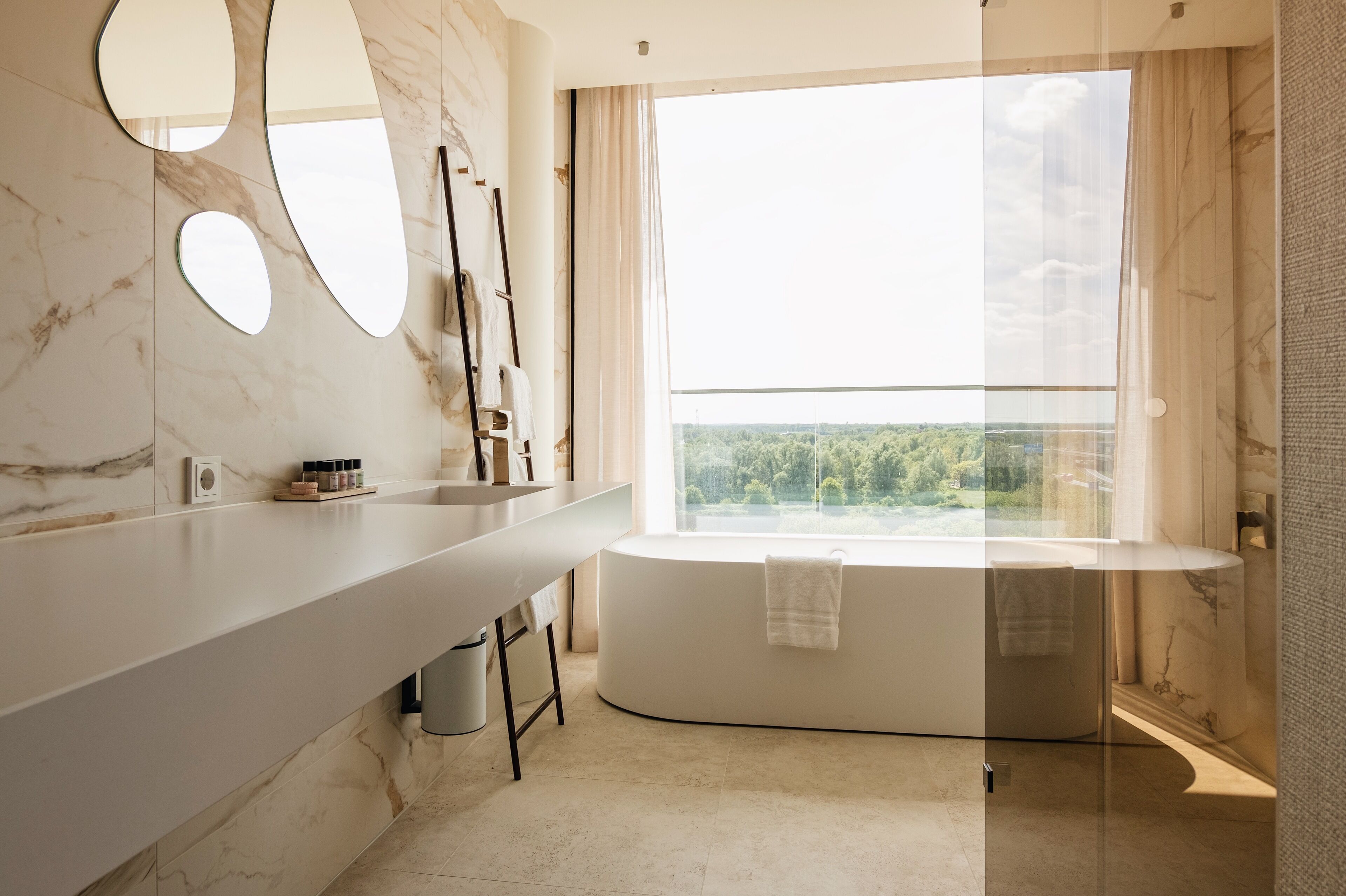 A modern bathroom with a marble vanity, a bathtub, and a large window offering a view of greenery.