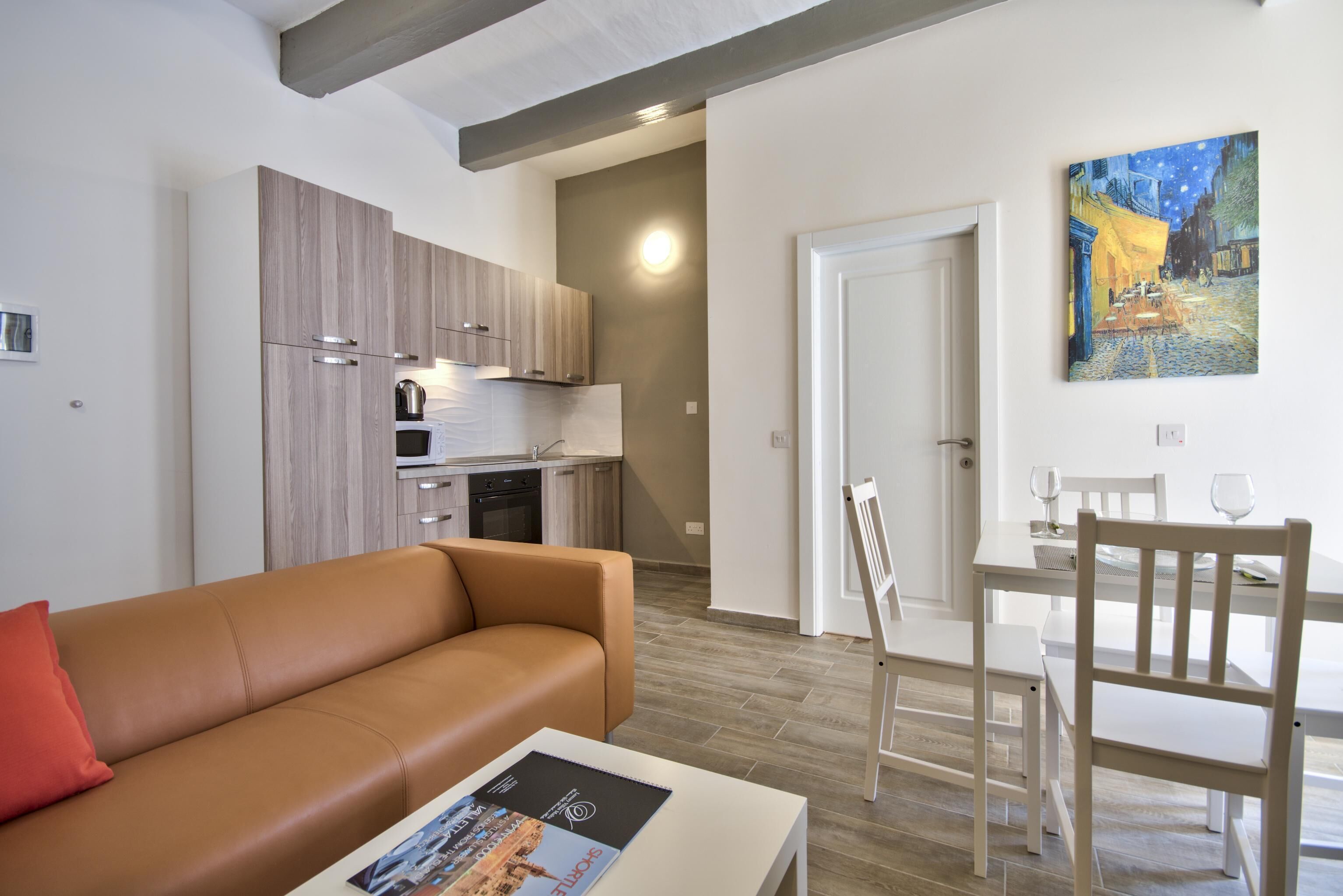 Een moderne woonkamer met een bruine bank, een witte salontafel met een tijdschrift, een eethoek met een witte tafel en stoelen, en een keuken met houten keukenkastjes en een schilderij aan de muur.