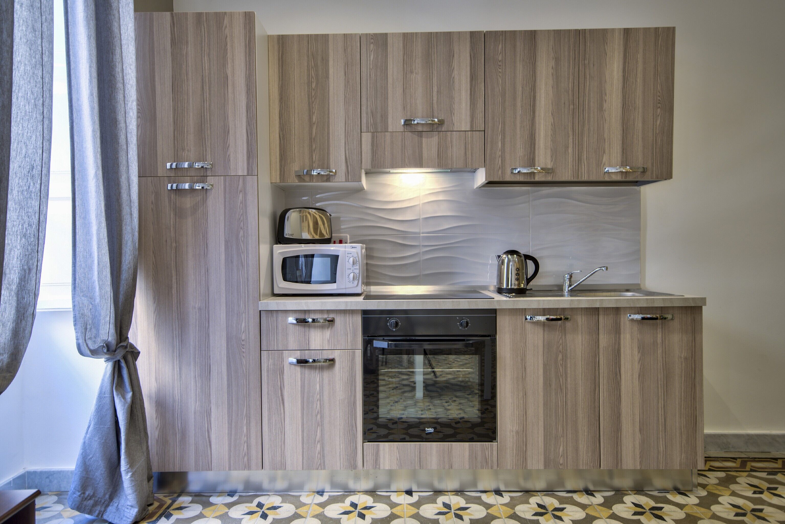 A compact kitchen with wooden cabinetry, a microwave, a kettle, and an oven.