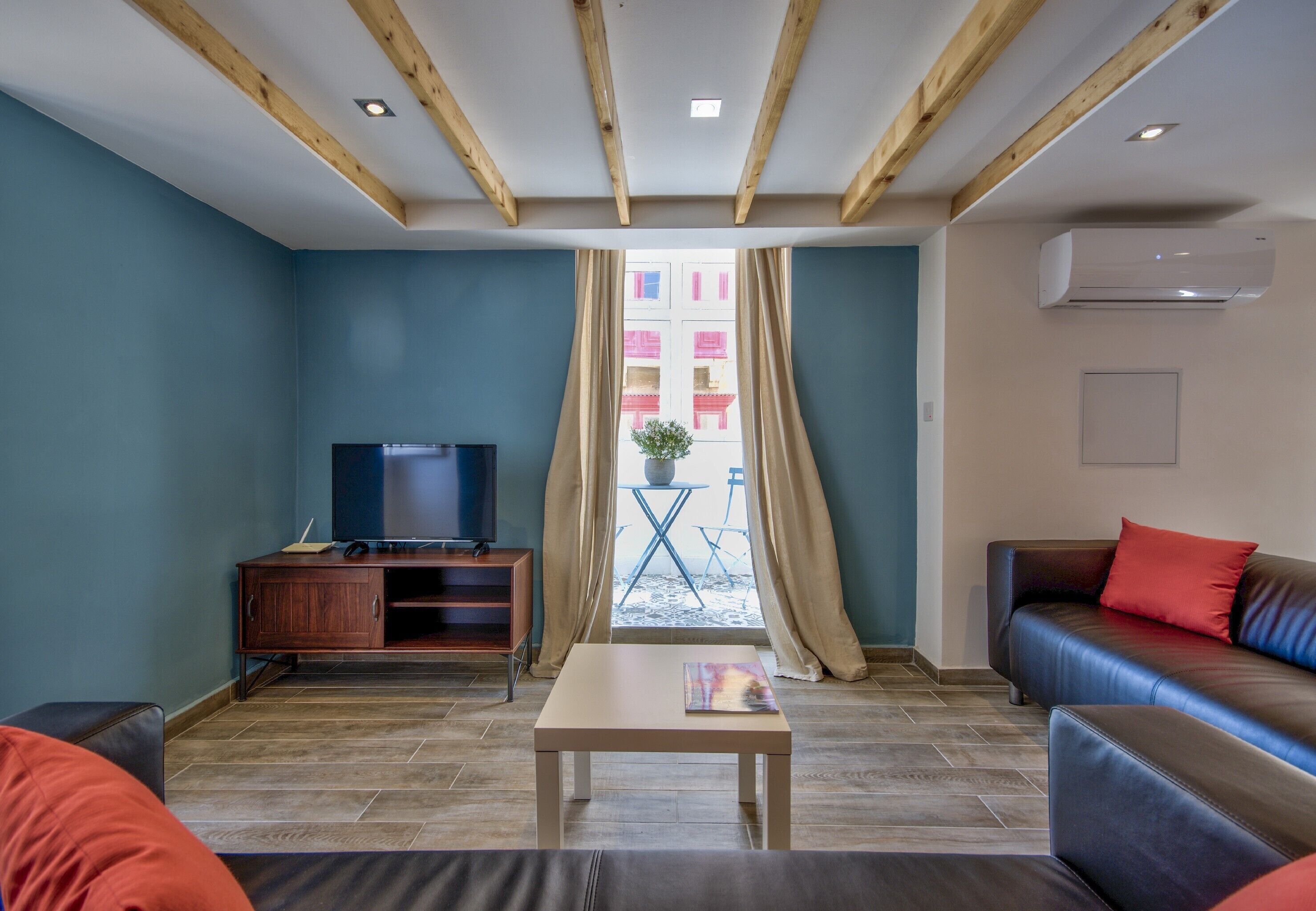 A modern living room with a wooden ceiling, a flat-screen TV on a wooden cabinet, a sofa with red cushions, and a table with a book.