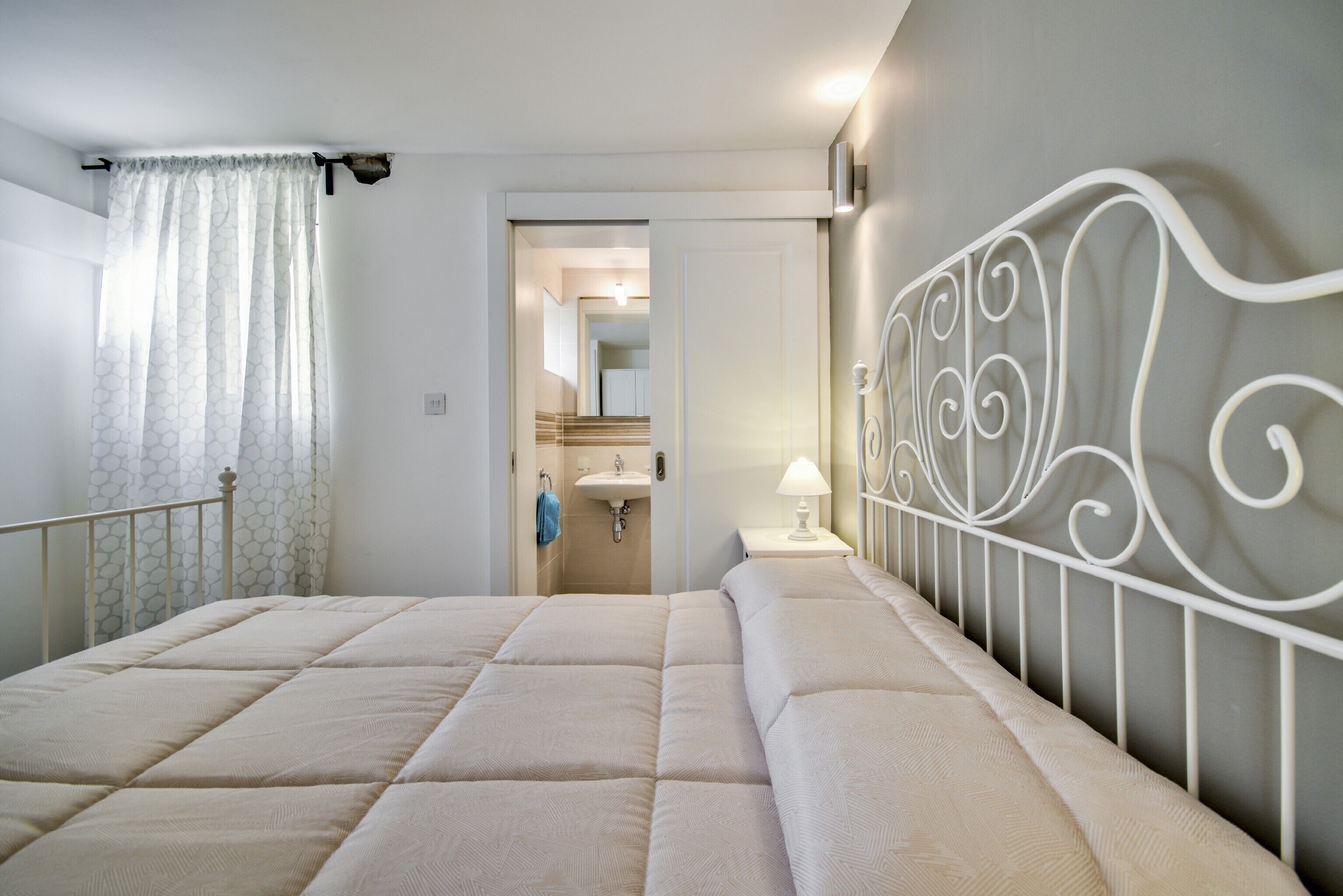A bedroom with a bed, a white metal headboard, a nightstand, a lamp, and a bathroom visible through an open door.