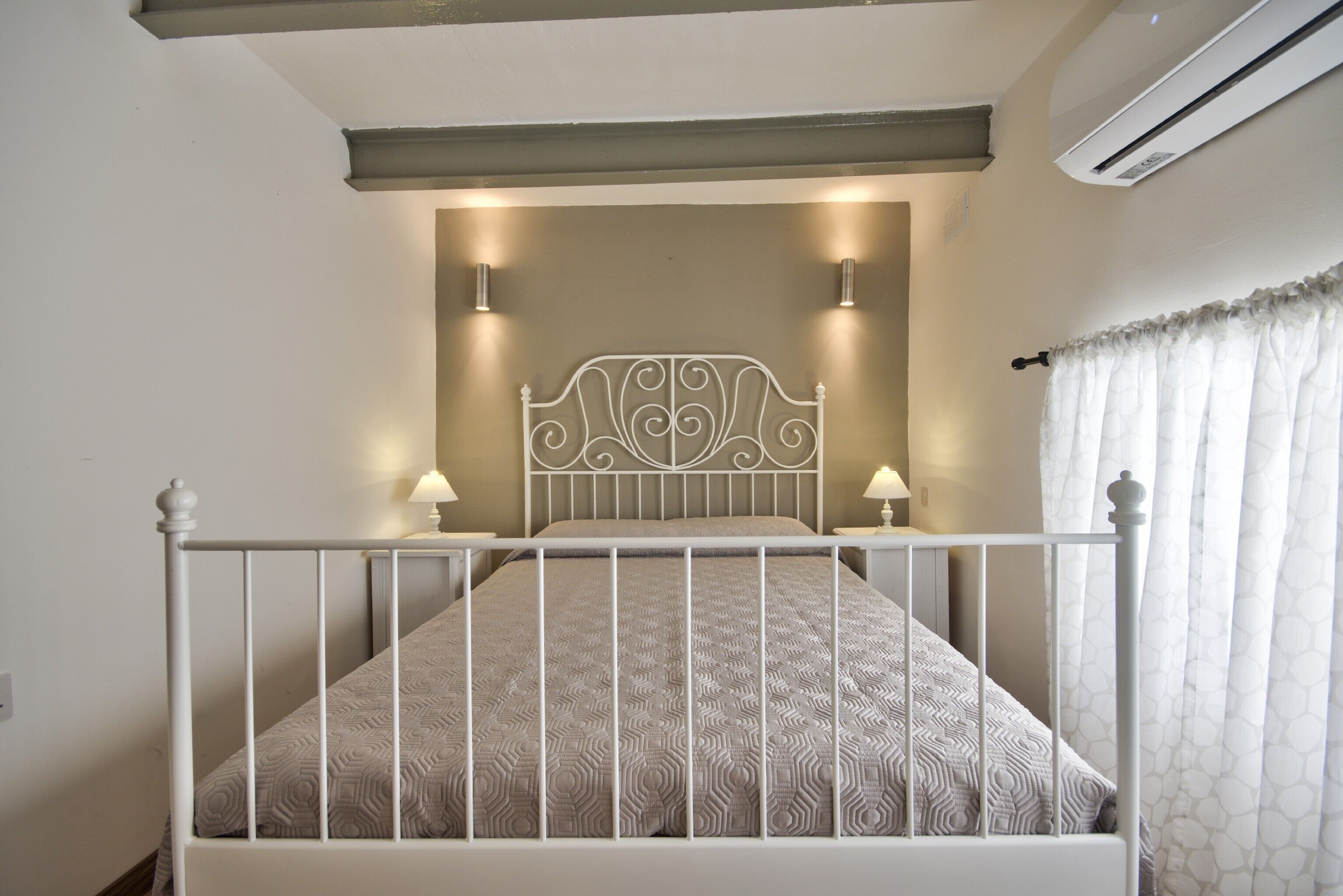 Una cama con estructura metálica blanca y ropa de cama estampada, dos lámparas de noche y un equipo de aire acondicionado arriba.