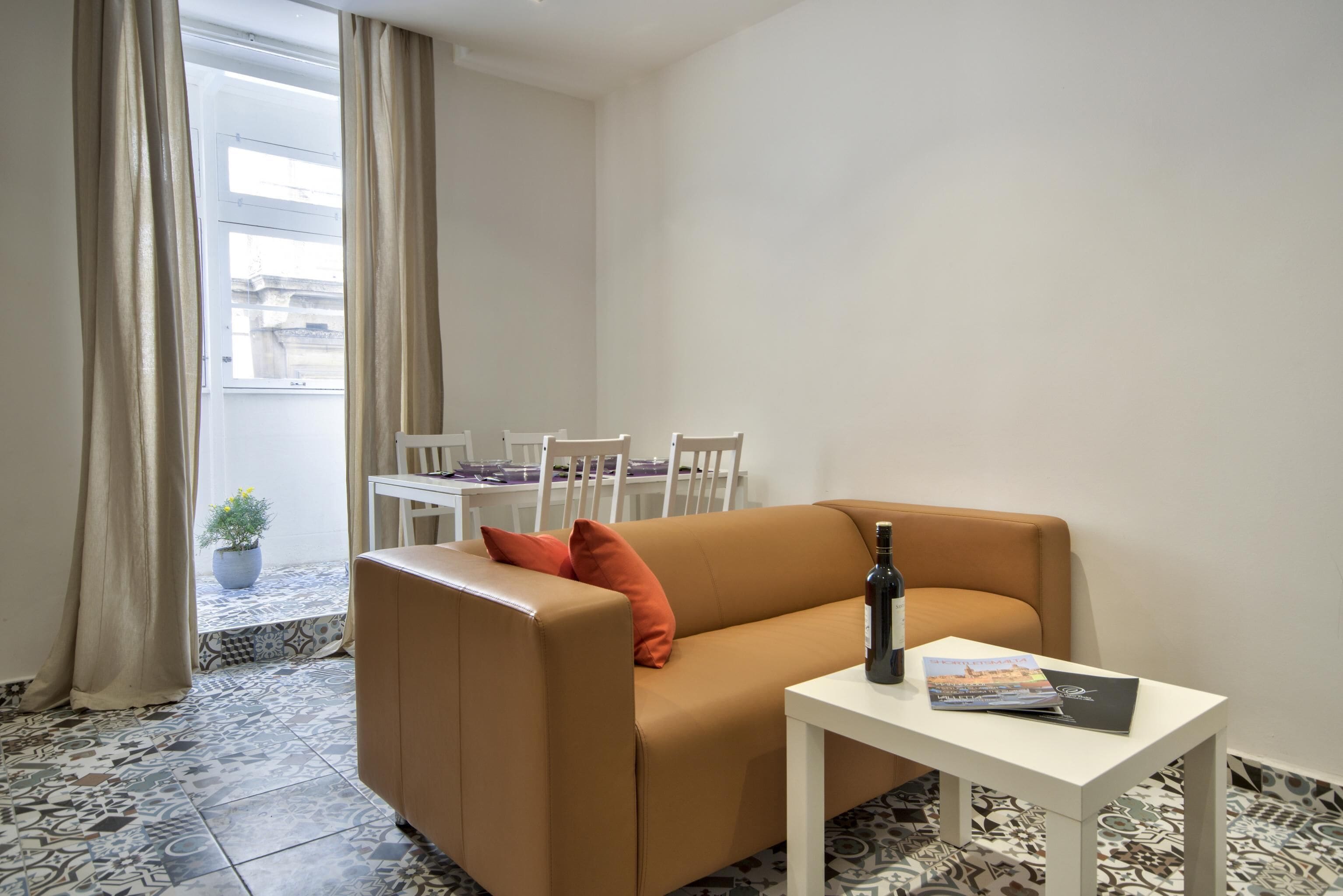 A modern living room with a brown sofa, a small white table, and a bottle of wine. A dining table with chairs is visible through a window with curtains.