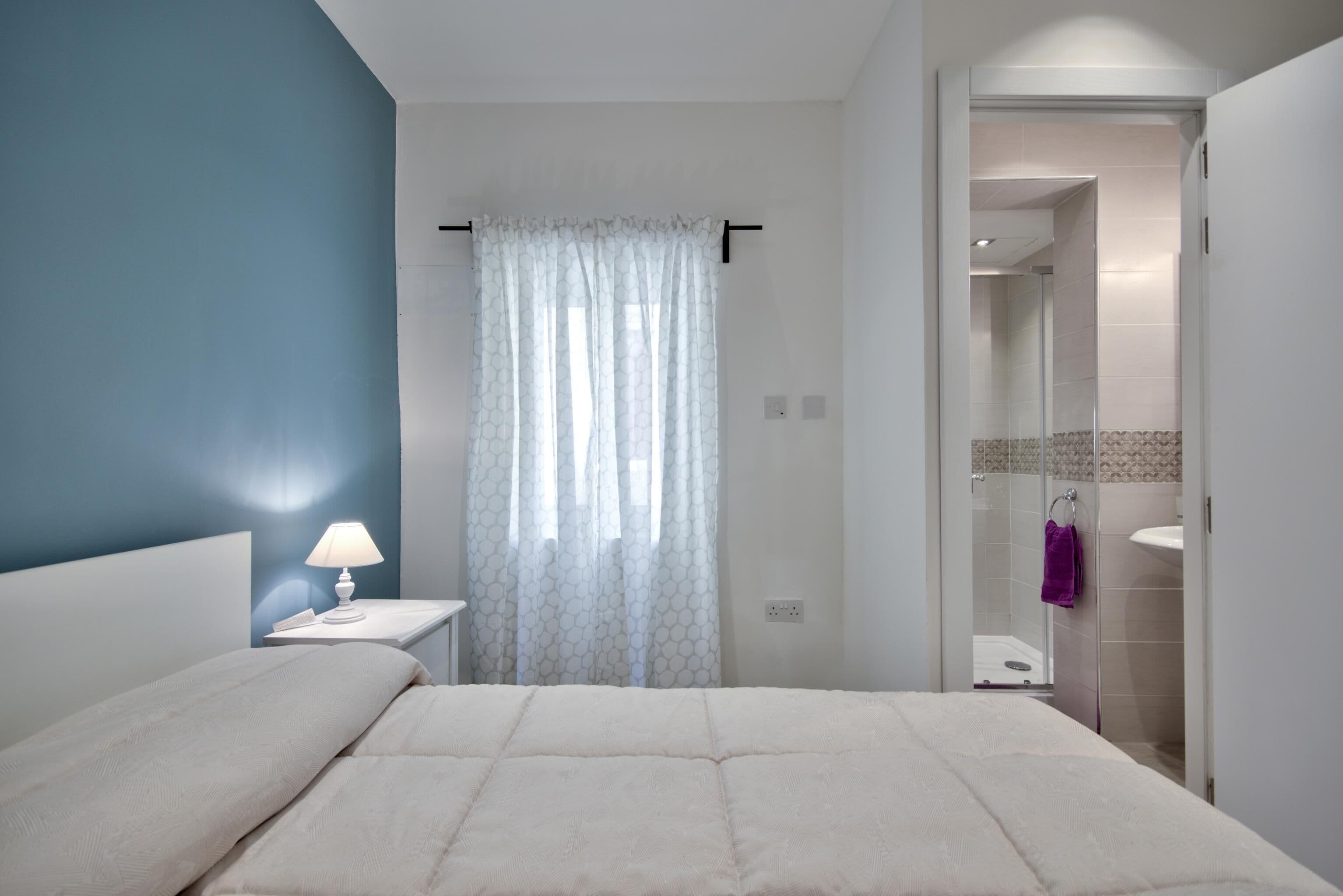 Un dormitorio con cama, mesita de noche, lámpara, baño con ducha y una toalla colgada en la puerta.