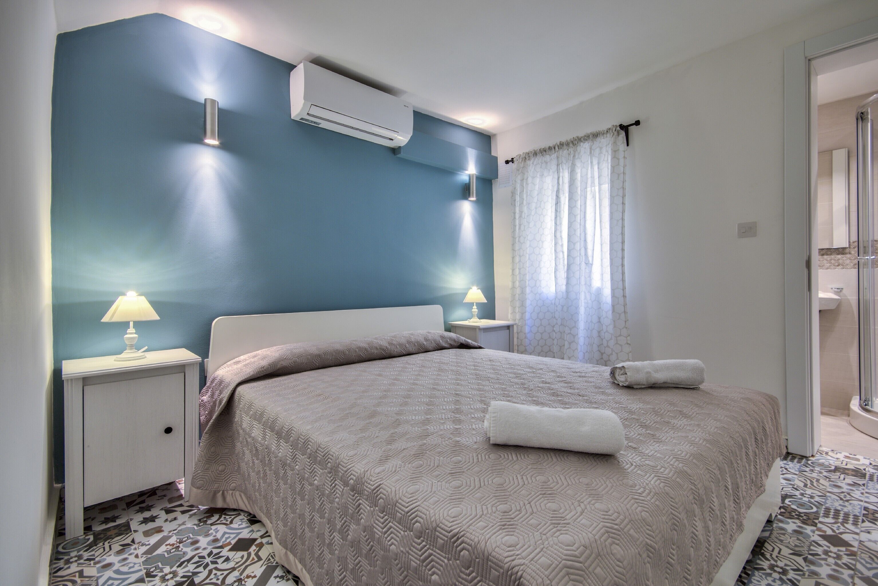 A bedroom with a bed, nightstands, an air conditioner, a window with curtains, and a bathroom visible through an open door.