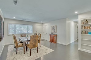 A spacious room with a dining area, a bookshelf, and a window with blinds.