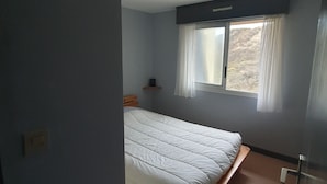 Ein Schlafzimmer mit einem Einzelbett, einem Fenster mit Vorhängen, einer kleinen Ablage und einer Tür.