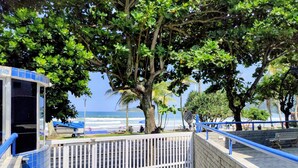 A coastal walkway with a metal gate, a tree-lined path, and a view of the ocean. - 01E Vista Linda Pe Areia Guaruja - Tombo (Guarujá)