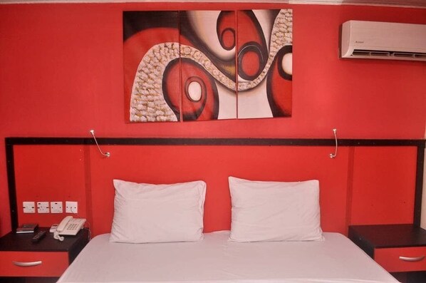 A hotel room with a red wall, a bed with white bedding, a painting above the bed, and an air conditioning unit on the wall.