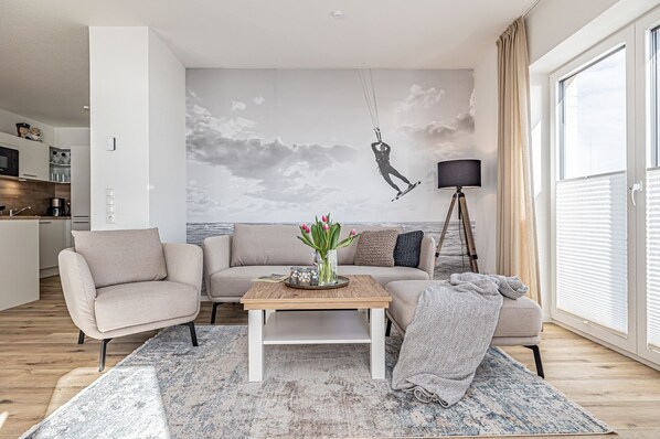 A modern living room with a sofa, coffee table, and a mural of a surfer. - Fjord-Koje 2 - Underfloor heating, shower, terrace, garden furniture,beach chair (Schleswig)