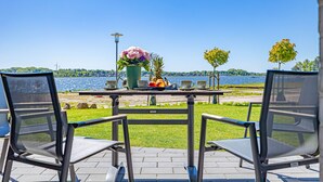 A table with chairs and a vase of flowers, overlooking a body of water with trees and a distant shoreline.