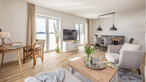Ein modernes Wohnzimmer mit Couch, Esstisch, Stühlen, einem Couchtisch und einem Fernseher. Große Fenster bieten Blick ins Freie.