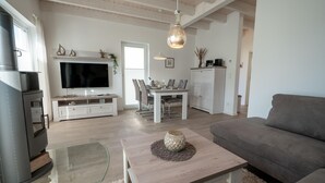 Una sala moderna con un televisor de pantalla plana, un comedor con una mesa y sillas blancas, un sofá gris y una mesa de centro de madera.