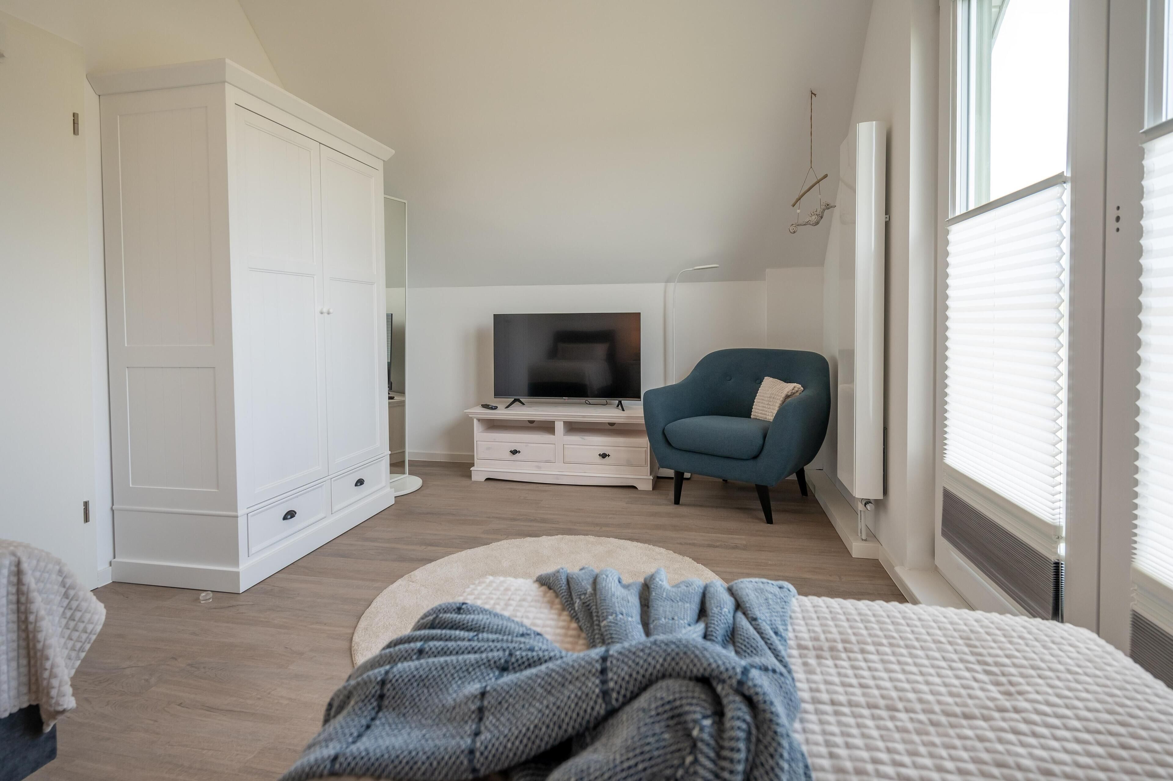 Een slaapkamer met een grote kledingkast, een televisie, een blauwe fauteuil en een groot raam met zonwering.
