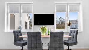 A modern dining area with a wooden table, four grey upholstered chairs, a flat-screen TV, and a vase with flowers.