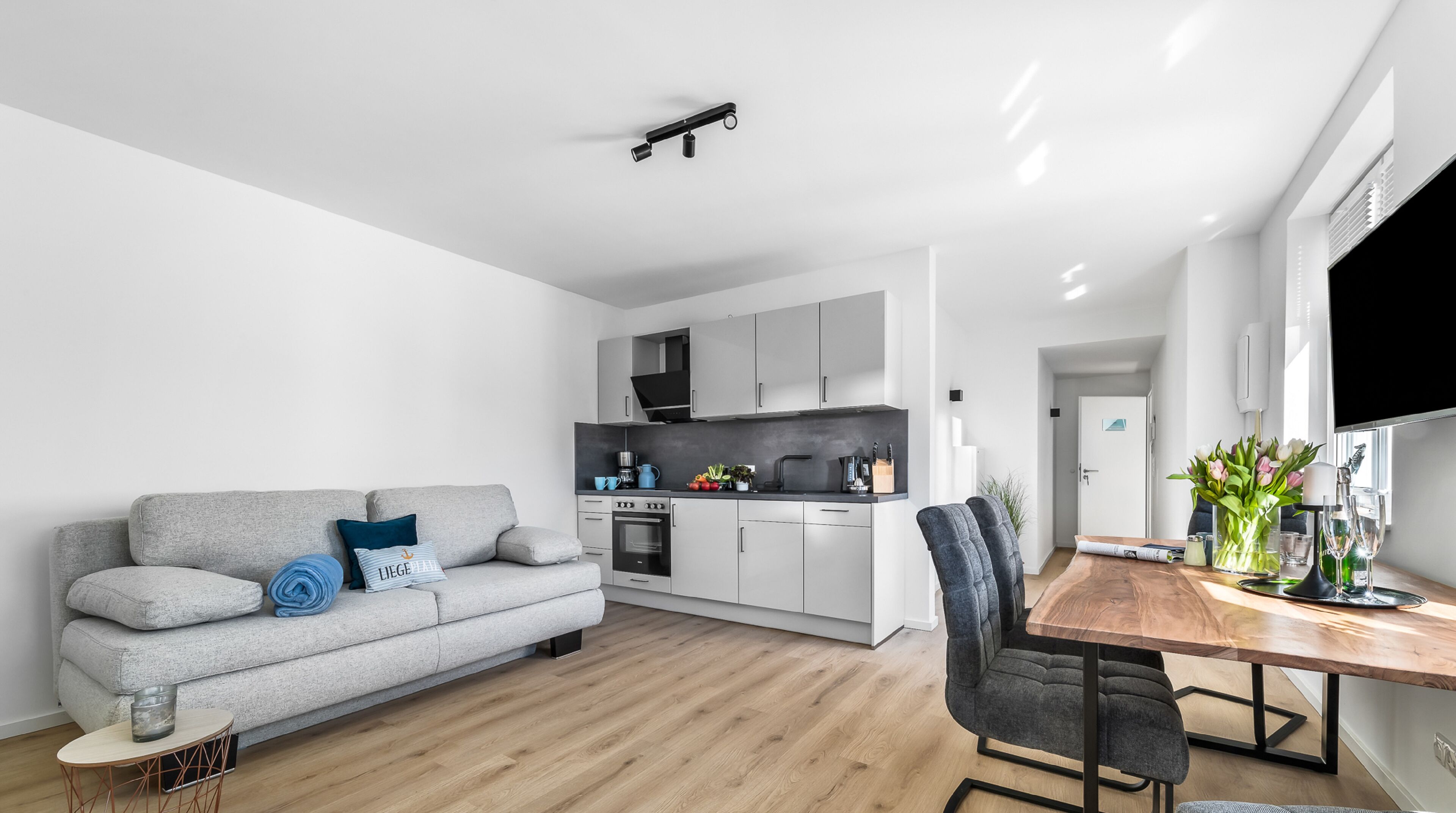 Um apartamento moderno com um sofá cinzento, uma mesa de jantar de madeira e uma área de cozinha.