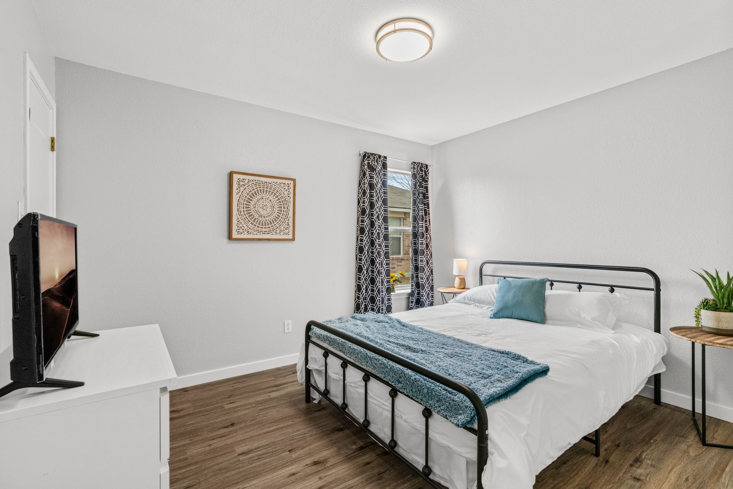 A bedroom with a bed, a TV, a bedside table, a lamp, and a window with patterned curtains.