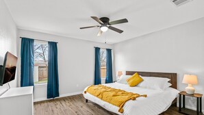 Un dormitori amb un llit, un ventilador al sostre, una finestra amb cortines, una taula de nit amb una làmpada i un sòl de fusta.