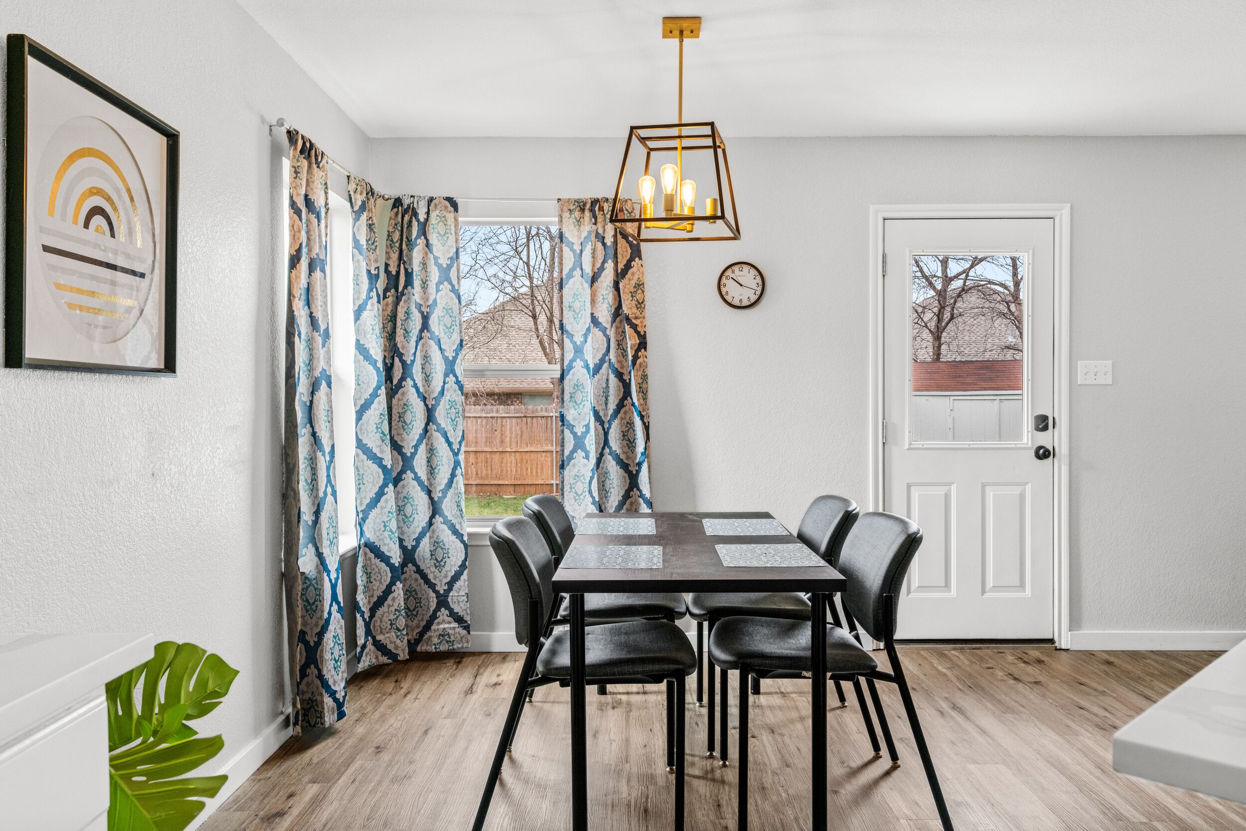 A dining area with a wooden table, black chairs, patterned curtains, a hanging light fixture, and a door with a view of trees.