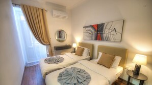 A hotel room with two beds, a round mirror, a wall art piece, and a small table with a lamp.
