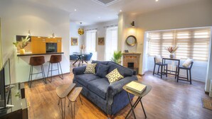 A modern living room with a blue sofa, wooden flooring, a kitchen area with bar stools, and a fireplace.