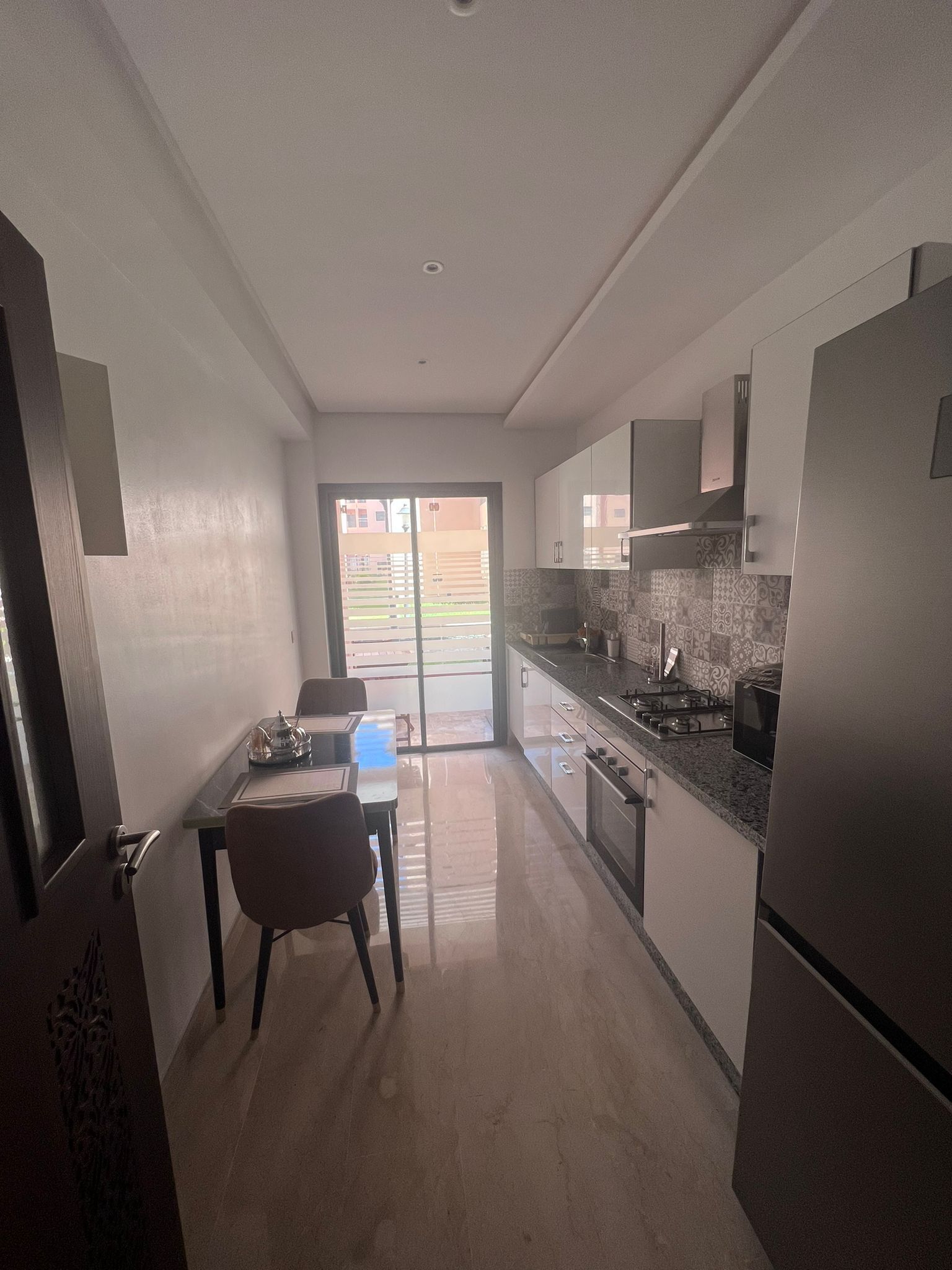 A modern kitchen with a dining area, a sliding glass door leading to a balcony, and a stainless steel refrigerator.