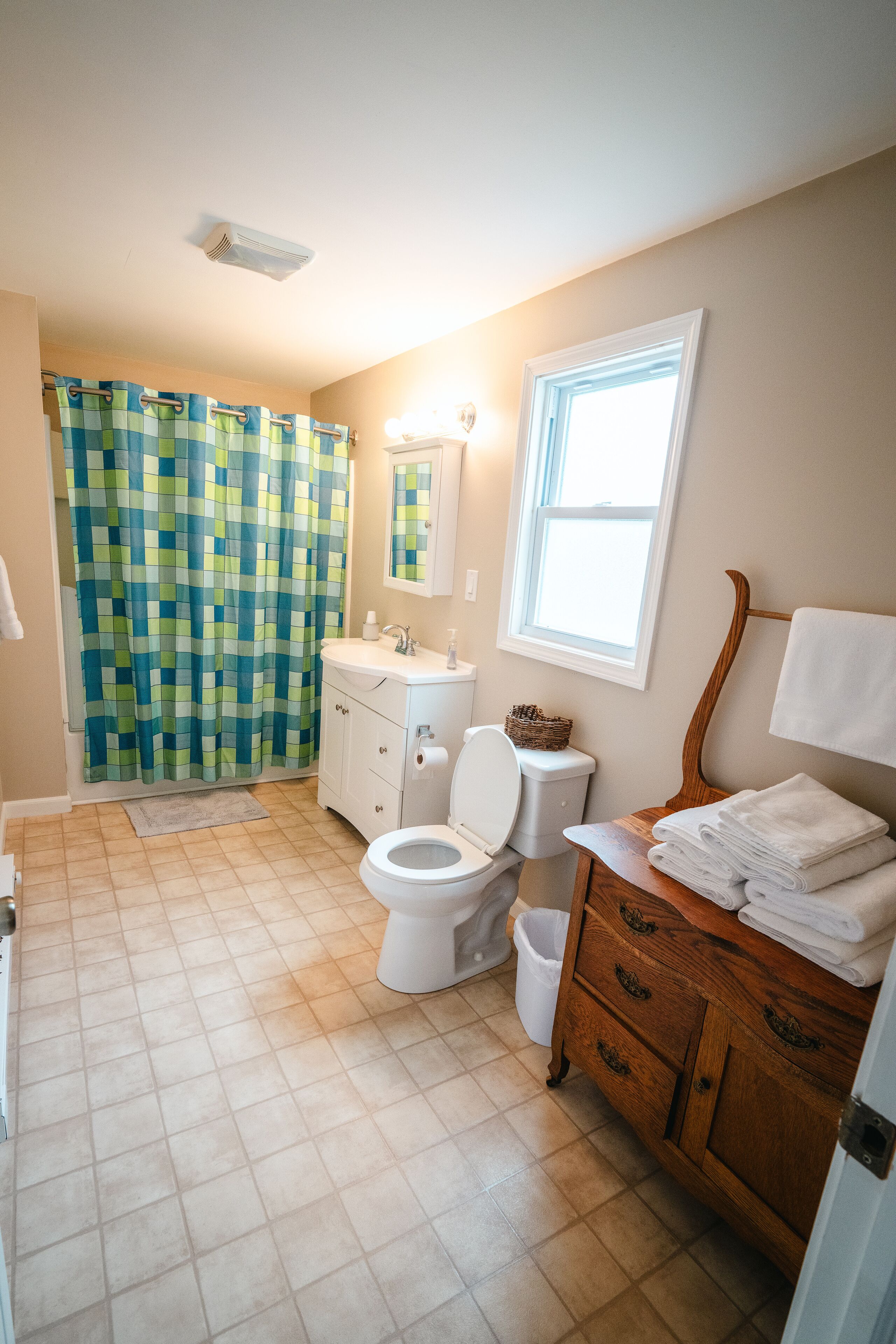 Une salle de bain avec un rideau de douche à carreaux, des toilettes blanches, un meuble-lavabo en bois avec un miroir et une fenêtre laissant entrer la lumière naturelle.