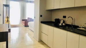 Une chambre d’hôtel moderne dotée d’un coin cuisine avec des meubles blancs, un plan de travail noir et un évier. On y trouve un lit avec une tête de lit blanche et une fenêtre avec des rideaux.