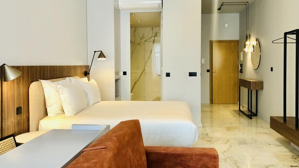 Une chambre d’hôtel moderne équipée d’un lit, d’un bureau, d’une chaise, d’un canapé, d’une lampe, d’un miroir et d’une salle de bain visible à travers une porte ouverte.