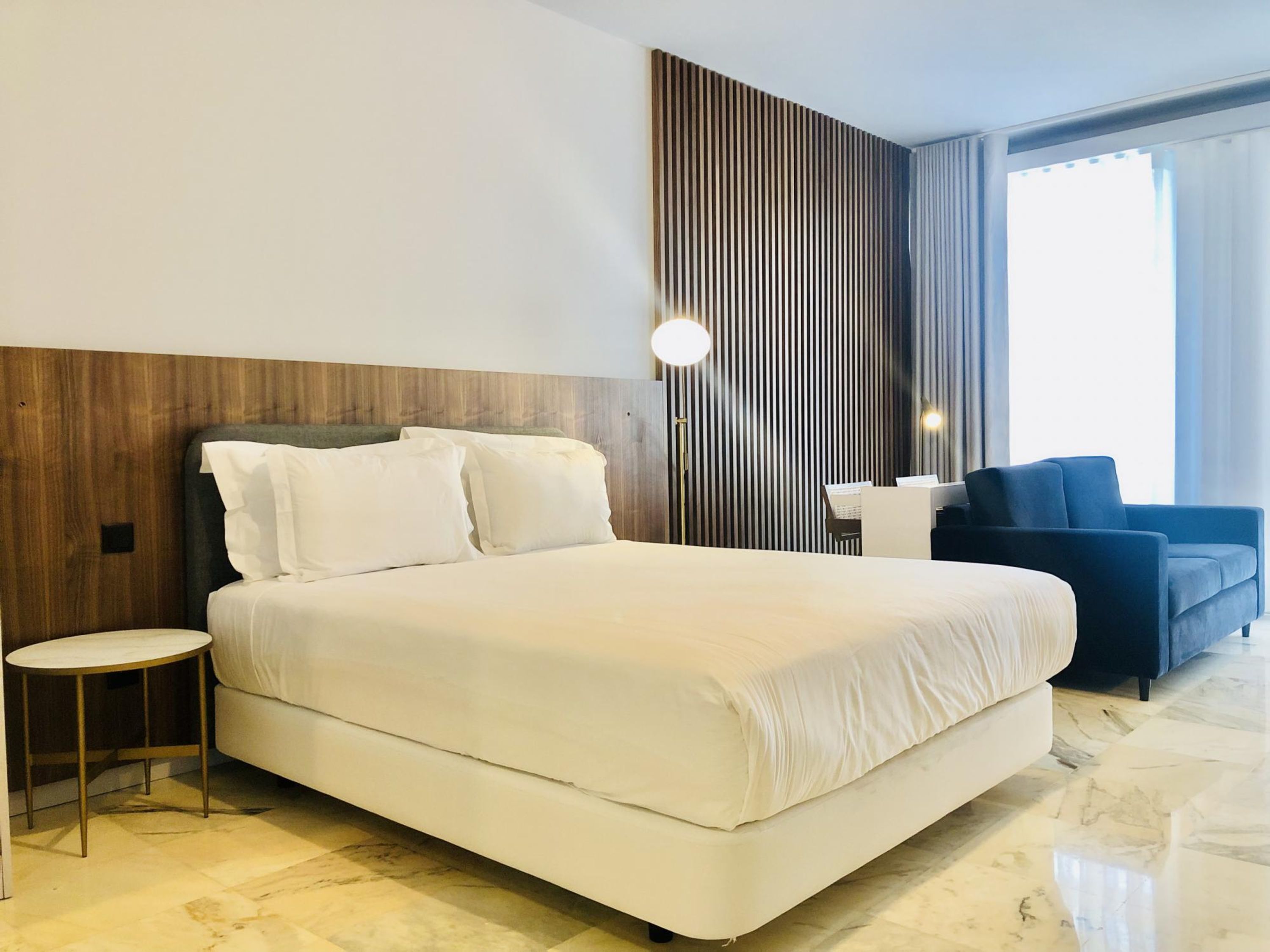 A modern hotel room with a large bed, a small side table, a desk with a chair, and a sofa.
