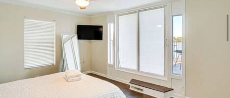 A bedroom with a bed, a ceiling fan, a TV mounted on the wall, and large windows.