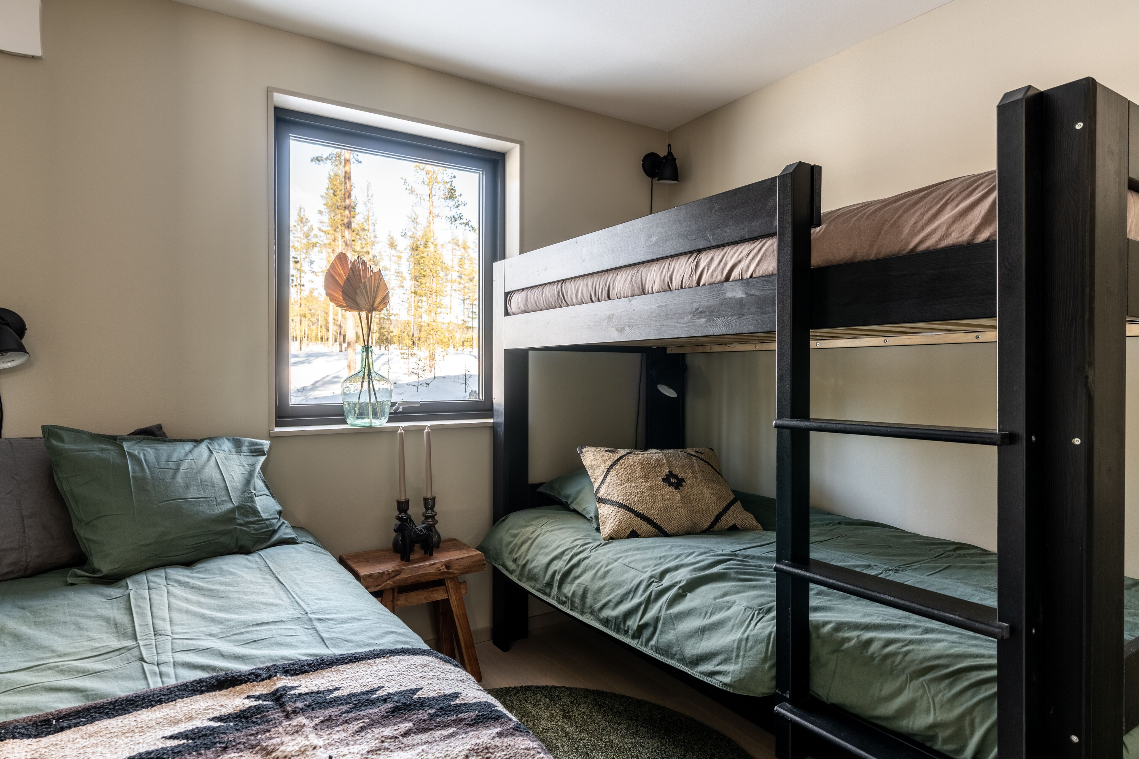A dormitory room with bunk beds, a window with a view of trees, and a bedside table with a lamp and a vase.