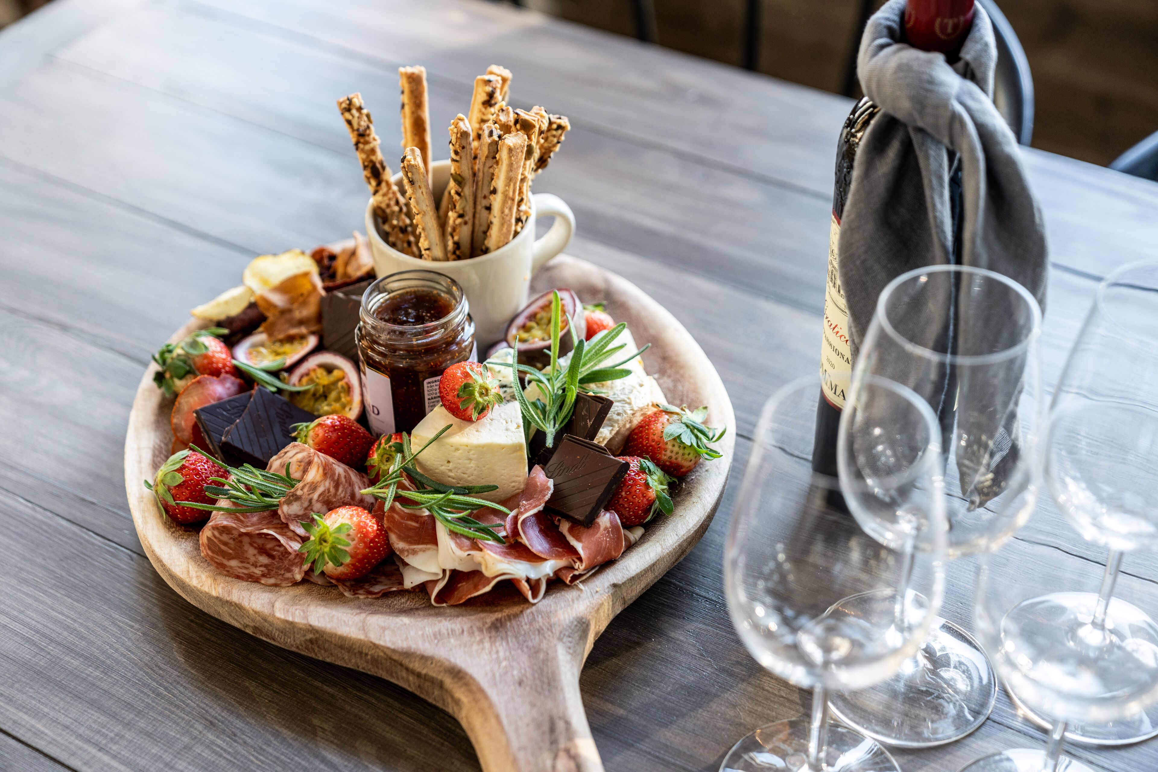 A wooden board with assorted cheeses, strawberries, and prosciutto, accompanied by crackers and a bottle of wine.