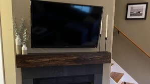 Ein modernes Wohnzimmer mit Flachbildfernseher über einem Kamin, Holzboden und einer Treppe im Hintergrund.
