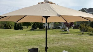 Eine Terrasse im Freien mit einem für vier Personen gedeckten Tisch, umgeben von weißen Stühlen und einem beigefarbenen Sonnenschirm.