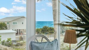 A view through a window with a palm tree, a chair, and a small table, overlooking a beach and a house.