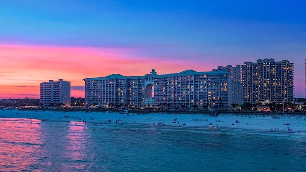A large hotel with multiple floors and a central courtyard, located near a beach with people on the sand and water.