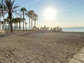 A beach with palm trees and a sunlit path leading to a sign that reads "Malagueta". - Hostal Moscatel (Málaga)
