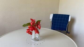 Una mesa redonda blanca con un jarrón de cristal transparente que contiene flores rojas. Un panel solar azul está montado sobre un soporte blanco frente a una pared lisa.