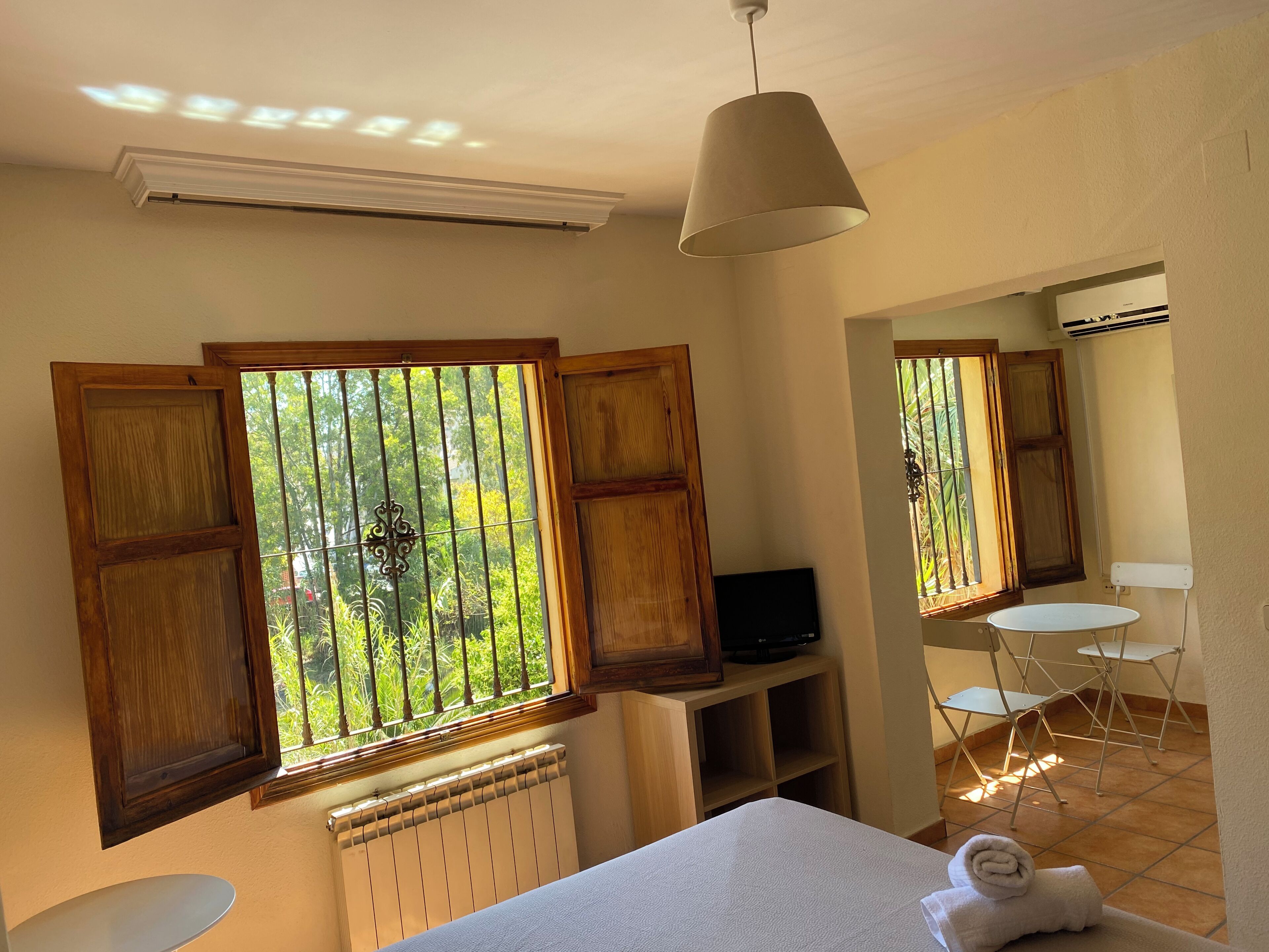 Una habitación con cama, televisor sobre un mueble, radiador, ventana con contraventanas de madera y una mesita con una toalla.