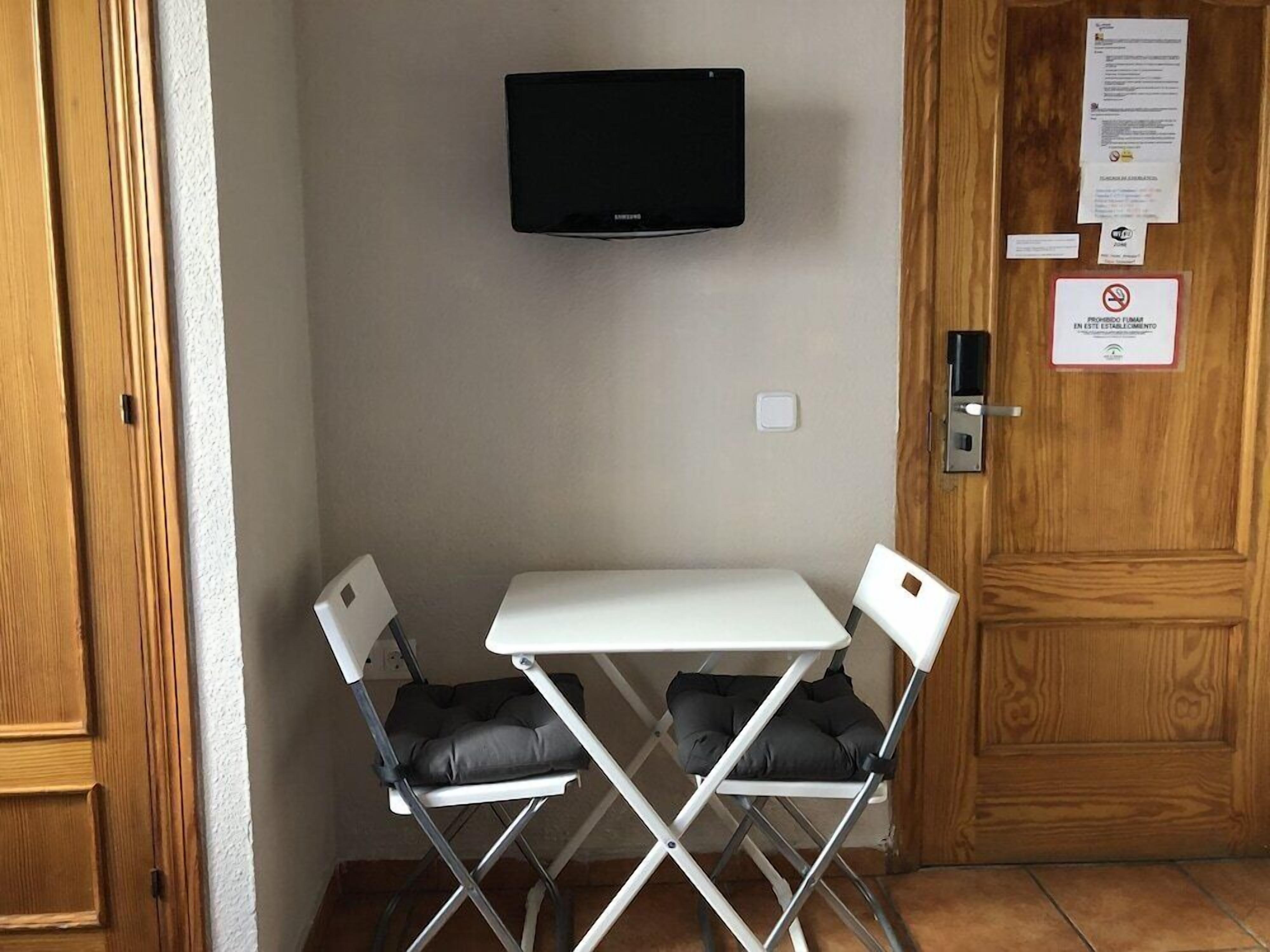 Una habitación pequeña con una mesa blanca y dos sillas, un televisor de pantalla plana montado en la pared, y una puerta de madera con un letrero.