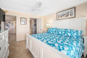 Une chambre à coucher comprenant un lit, un ventilateur de plafond, une commode, une table de chevet et un tableau encadré accroché au mur.
