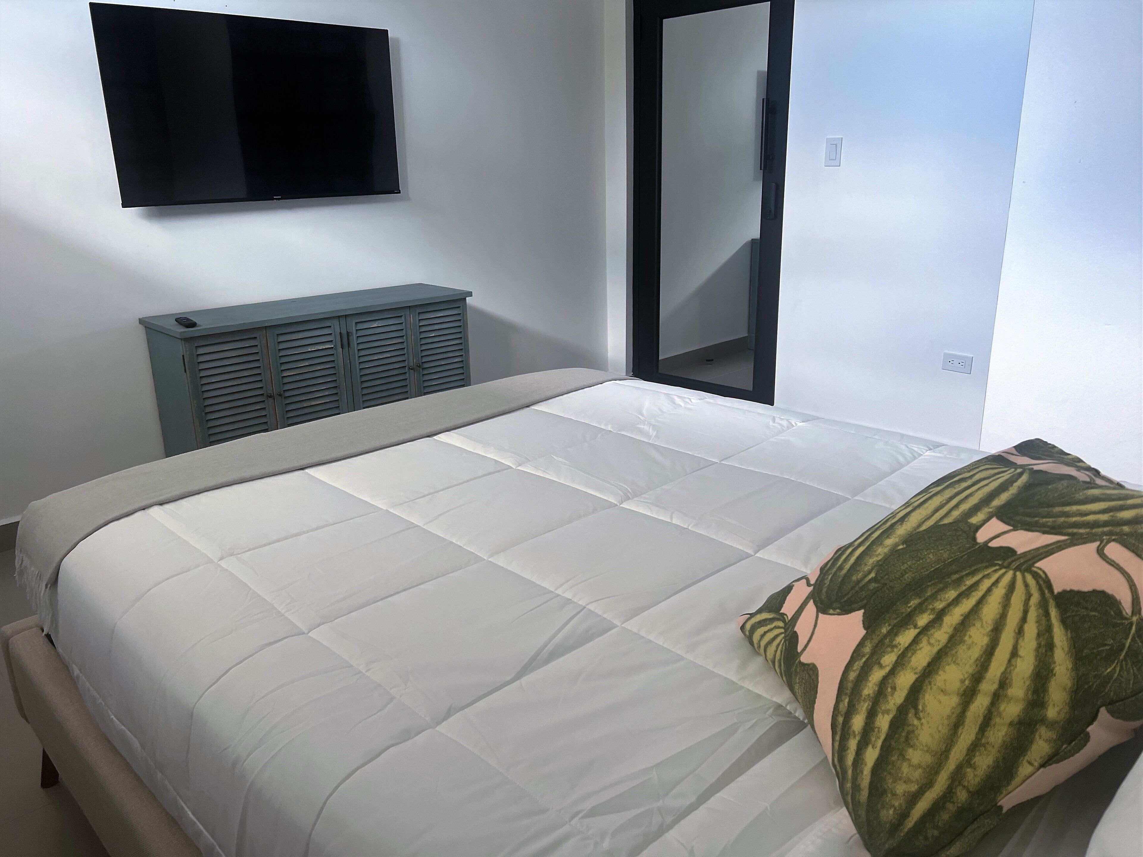 A neatly made bed with a white duvet and a single decorative pillow, a gray cabinet with a remote control on top, and a flat-screen TV mounted on the wall.