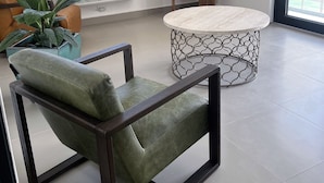 Una sala de estar moderna con un sillón de cuero verde, una mesa de centro redonda con base de metal geométrico, una estantería de madera y un amplio ventanal que ofrece vistas de edificios.
