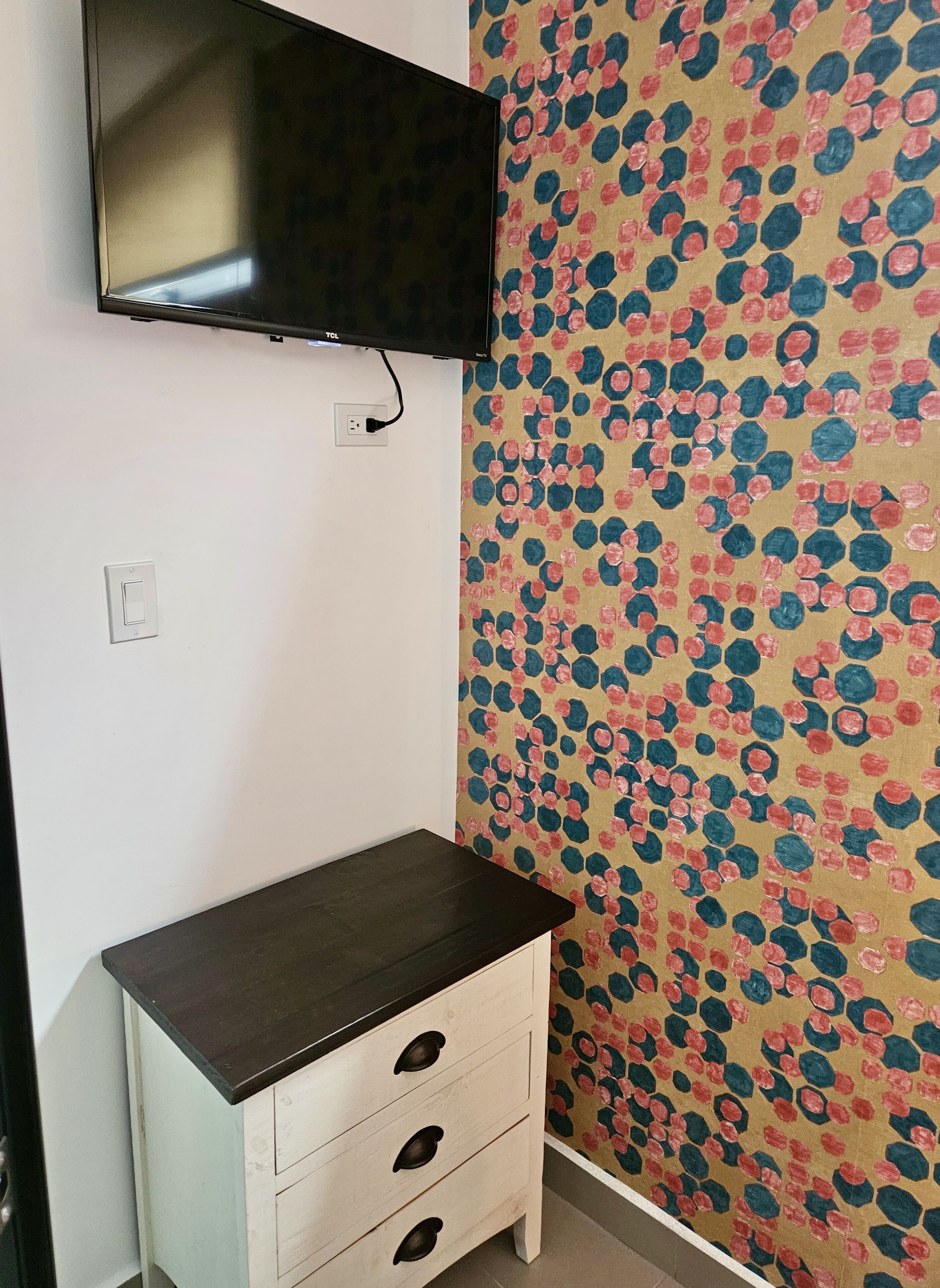 A room with a flat-screen TV mounted on the wall, a small white dresser with drawers, and a patterned wallpaper.